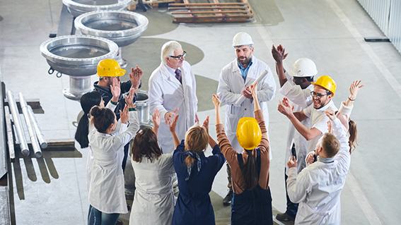Gruppe von Mitarbeitenden in Arbeitskleidung und Schutzhelmen steht in einer Produktionshalle im Kreis und klatscht gemeinsam. Die Szene zeigt Job Rotation als Teil der Zusammenarbeit und des Wissensaustauschs im Team. Gruppe von Mitarbeitenden in Arbeitskleidung und Schutzhelmen steht in einer Produktionshalle im Kreis und klatscht gemeinsam. Die Szene zeigt Job Rotation als Teil der Zusammenarbeit und des Wissensaustauschs im Team.